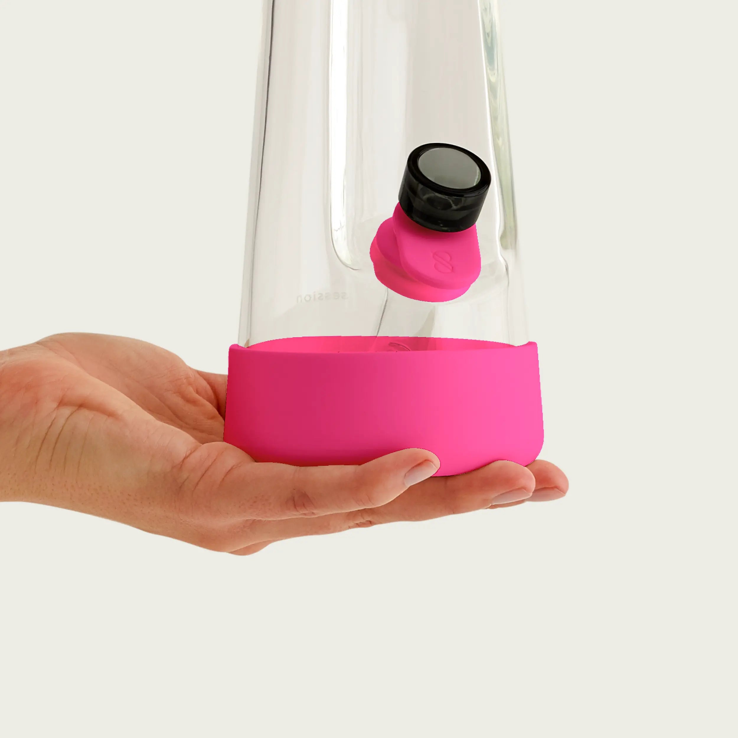 A hand holding the base of a clear glass Session Bong fitted with the Magenta Pink silicone accessory set. The image showcases the snug fit of the pink protective footer and the pink pull tab attached to the glass bowl.