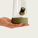 A close-up shot of a hand supporting the base of a Session Goods bong, highlighting the olive green silicone footer. The image shows the protective silicone sleeve fitted to the bottom of the tapered glass and a matching olive tab on the bowl.