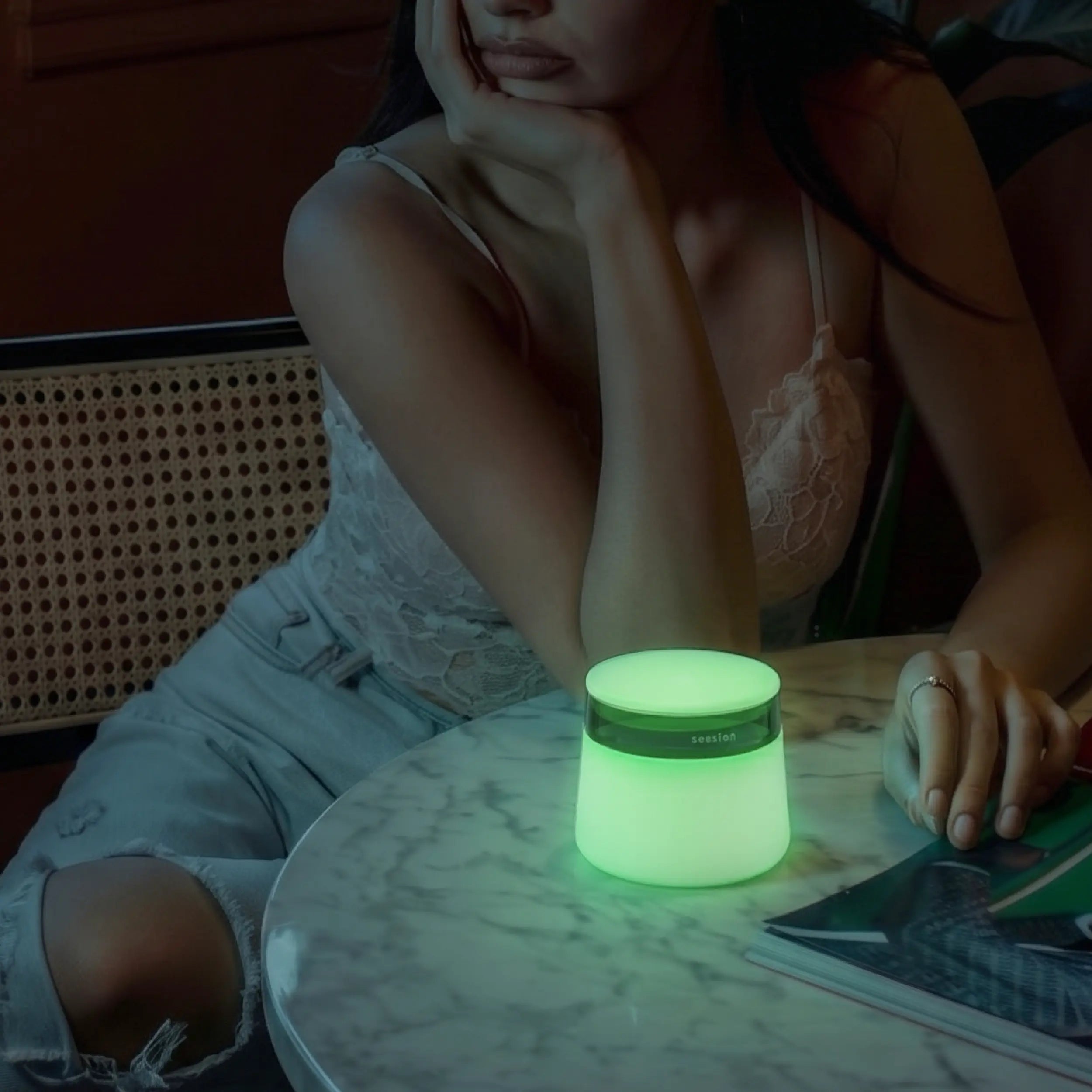 A woman sits at a marble table in a dimly lit room next to a fully illuminated, glow-in-the-dark lime green Session Goods stash jar and ashtray.