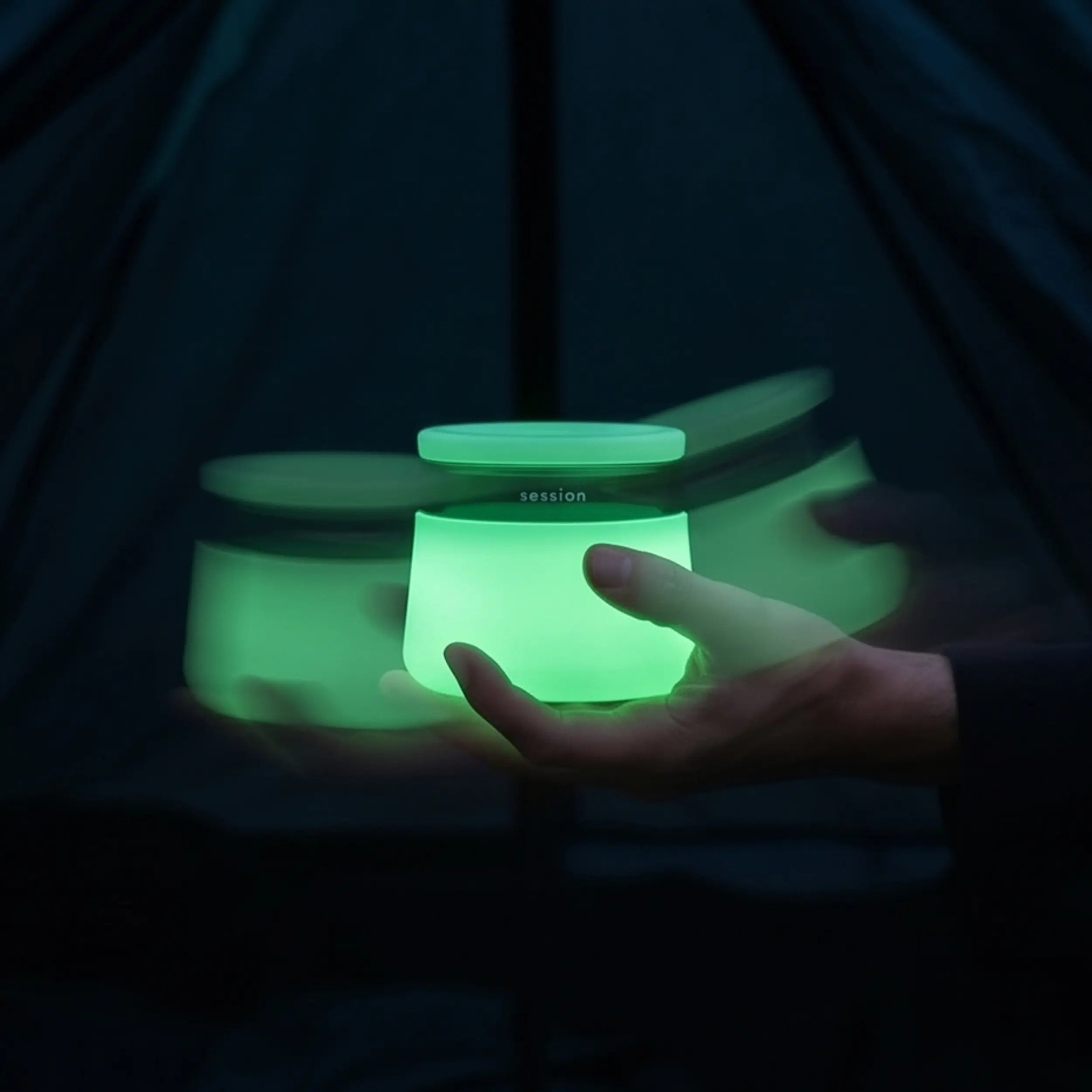 Hand holding a brightly glowing lime green Session Goods stash jar and ashtray in a dark, outdoor setting, featuring a stylized motion blur effect.